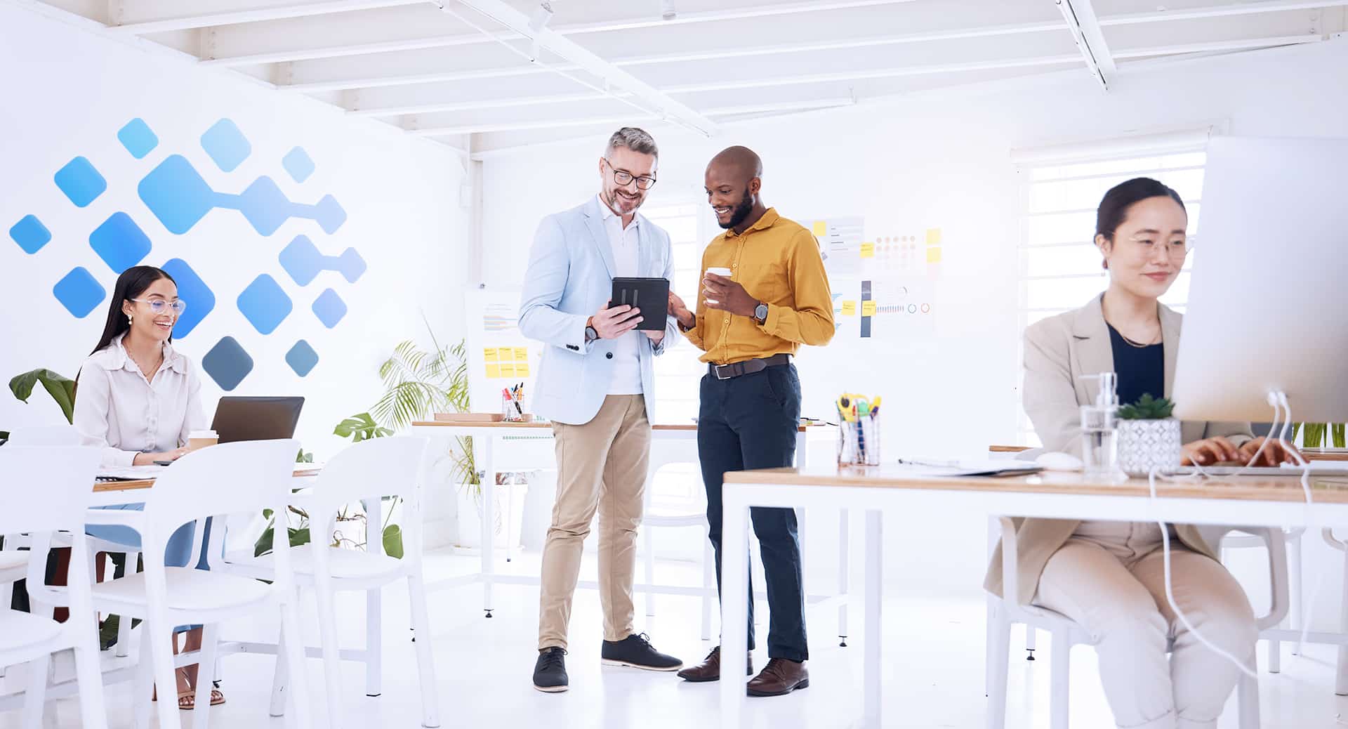 Office environment with professionals discussing iMIS software upgrades, one man holding a tablet, another man with a coffee, woman working on a laptop, and blue geometric wall design in background.